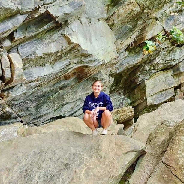 Person wearing a sweatshirt, crouching on rocks in front of a rocky cave wall.