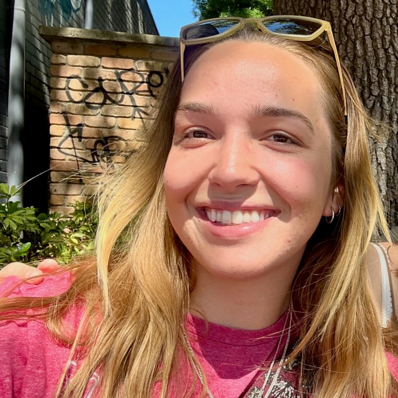 Smiling person with sunglasses on head, brick wall and tree in background.